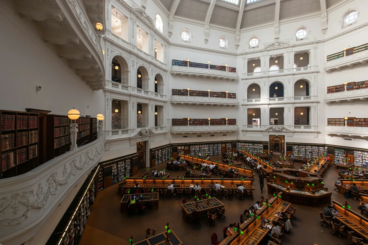 A library reading room with study desks and tiers of bookshelves.